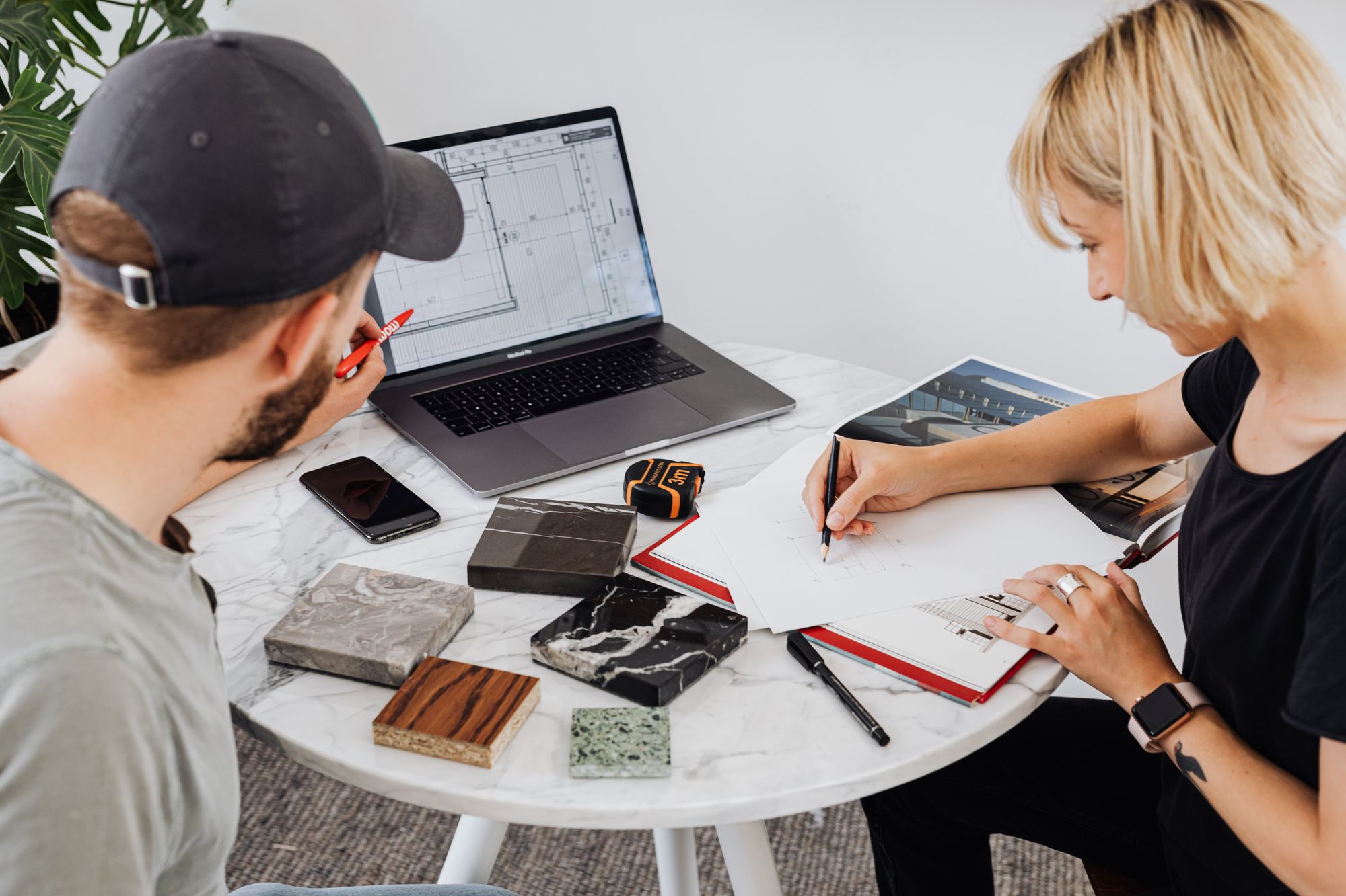 Two designers work with floor plans, a laptop, and material samples at a round table.