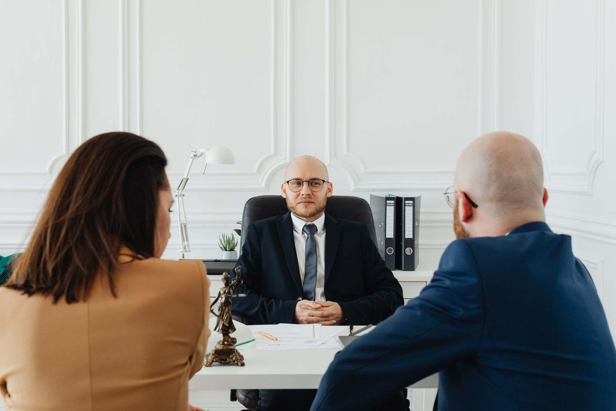 A lawyer meets with two clients across a desk in a bright office.