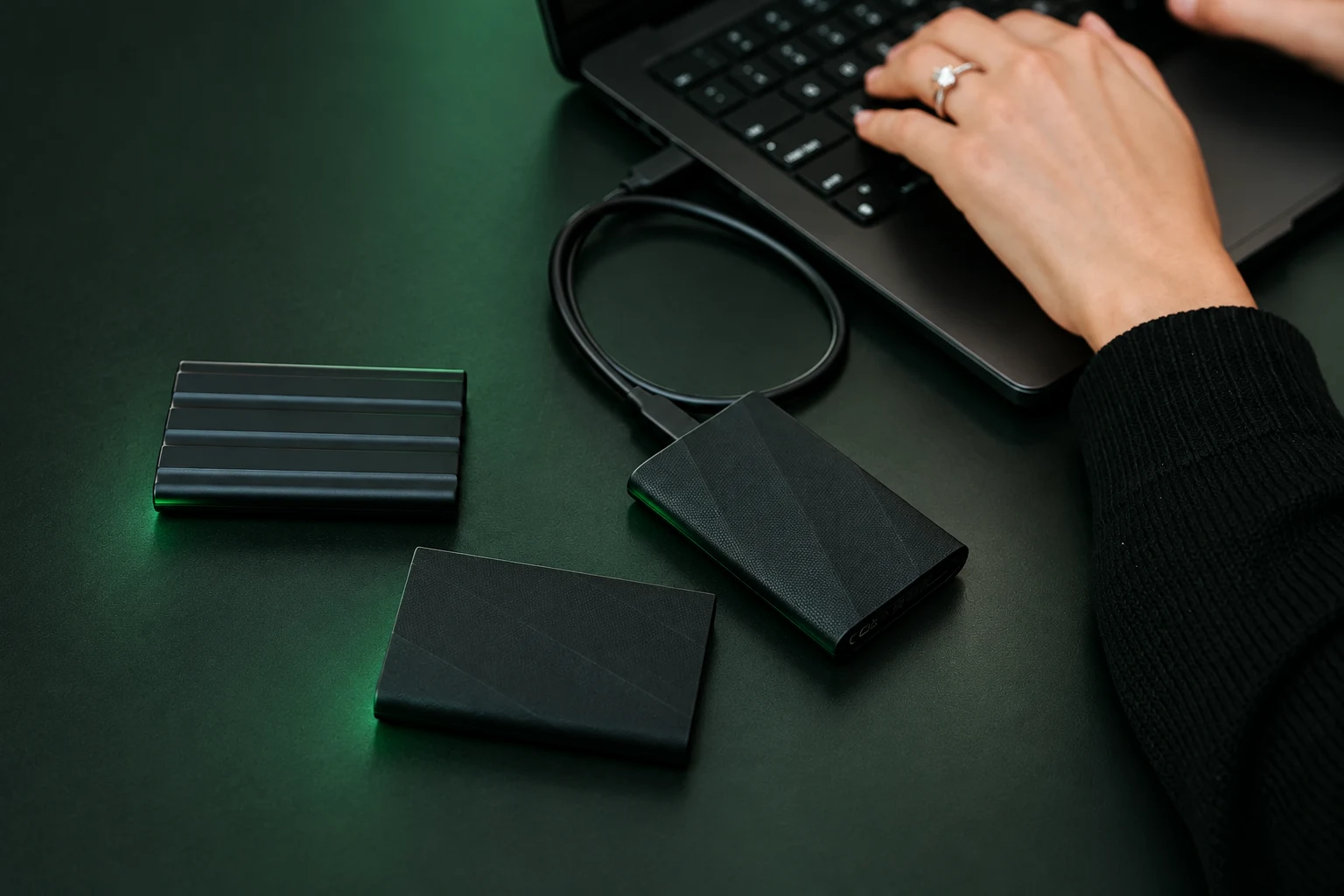 Black portable storage drives connected to a laptop on a dark-green work surface